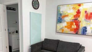 A modern room with a grey sectional sofa, a large abstract painting, a clock, and a door leading to a bathroom. - Cozy 1-bedroom cottage in serene Ravenel with AC (Ravenel)