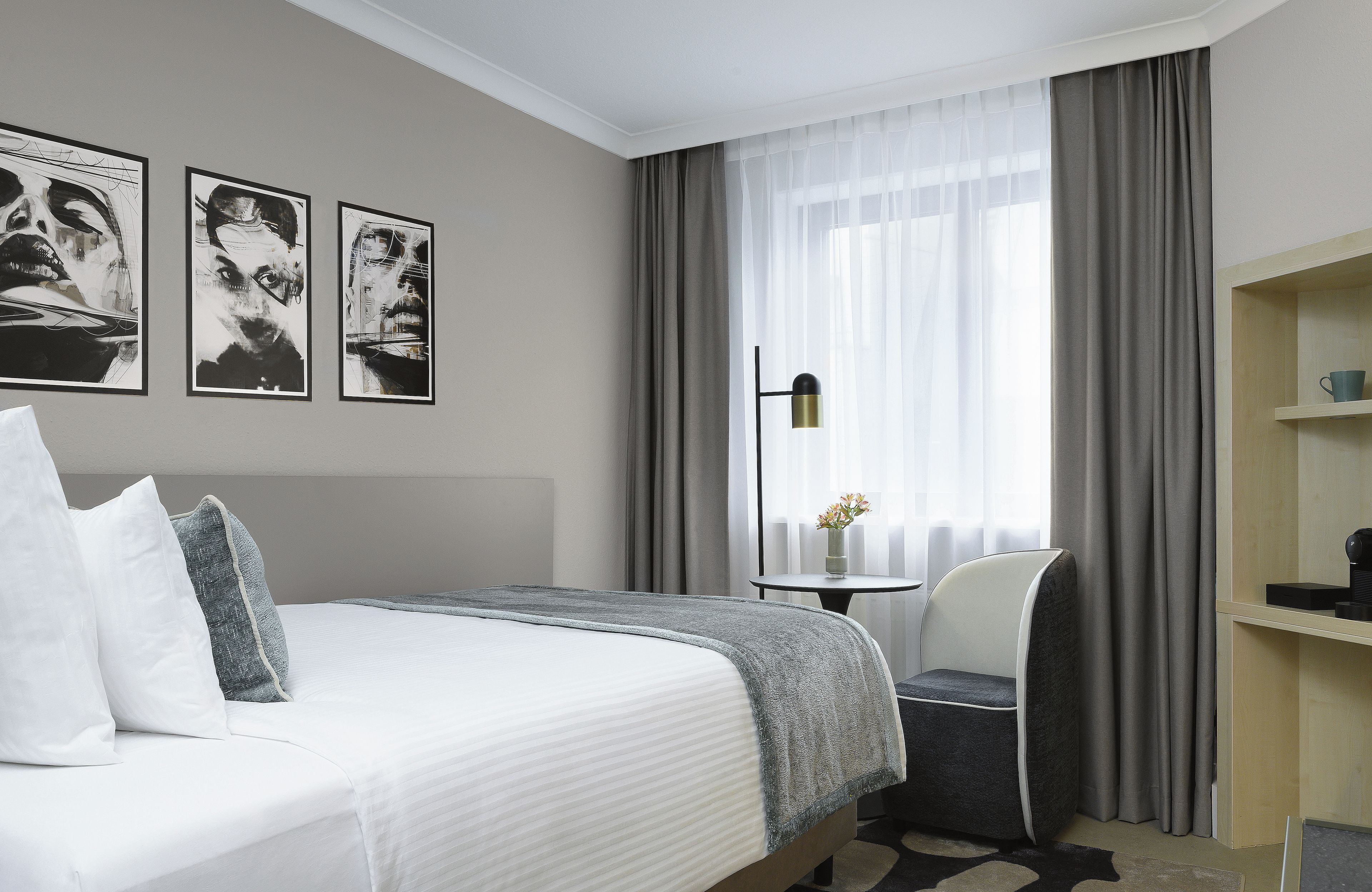 A hotel room with a bed, a chair, a small table with a vase, and a shelf with a cup.
