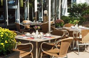 Mobiliario de cafetería al aire libre con sillas y mesas de mimbre, algunas con tazas y platos, y plantas en macetas.