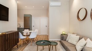 En moderne stue med sofa, spisebord med stole, et køkkenområde med et køleskab og et fladskærms-tv ophængt på væggen.