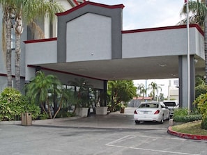 Ingresso di un hotel con vialetto coperto, palme e un'auto bianca parcheggiata all'interno.