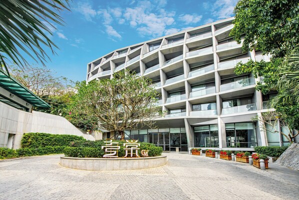 A modern building with a curved facade, a landscaped courtyard with a central tree, and a sign with Chinese characters. - CM+ Serviced Apartment Shenzhen Hillside (Shenzhen)