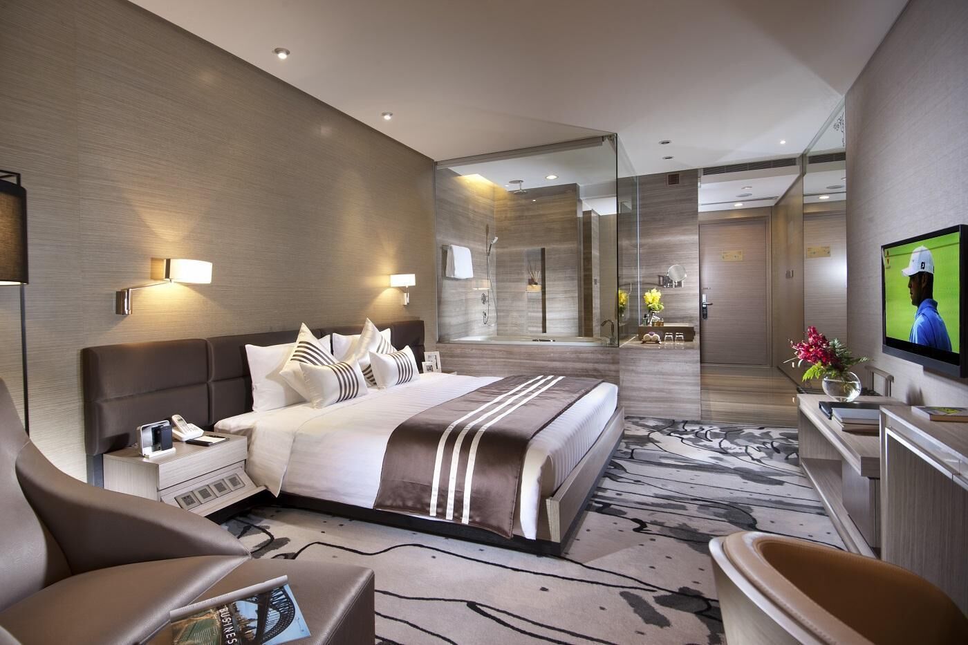 A modern hotel room with a large bed, a TV, a desk with a chair, and a bathroom visible through a glass partition.