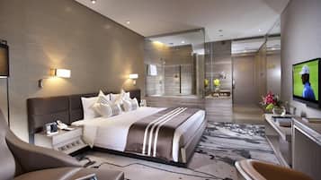 A modern hotel room with a large bed, a TV, a desk with a chair, and a bathroom visible through a glass partition.