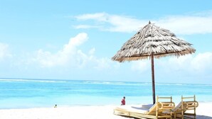 A beach scene with a straw umbrella, two wooden lounge chairs, and a person in a red outfit. - Diani Reef Beach Resort and Spa (Diani Beach)