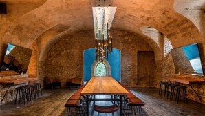 A long wooden table with multiple chairs in a rustic stone-walled room with a high ceiling and a hanging light fixture.