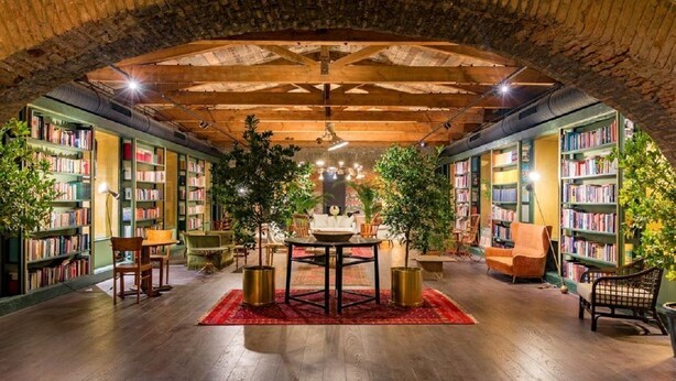 An indoor library with bookshelves, a central table, chairs, and potted plants.