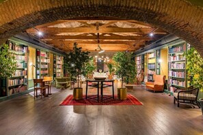 Una biblioteca interior con estanterías, una mesa central, sillas y plantas en macetas.