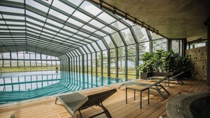 A covered outdoor swimming pool with wooden decking, lounge chairs, and potted plants.