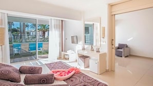 A hotel room with a bed, a desk, a chair, a sofa, a small table with a pink floral arrangement, and a sliding glass door leading to a pool area. - Dome Beach Marina Hotel and Resort (Ayia Napa)