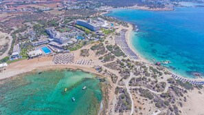 An aerial view of a coastal resort with a large hotel, multiple swimming pools, and a long beach with umbrellas. - Dome Beach Marina Hotel and Resort (Ayia Napa)