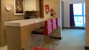 A modern kitchen with a white countertop, a microwave, and a fire extinguisher. There are four red bar stools and a painting on the wall. - Adagio Fujairah (Al-Fujairah)