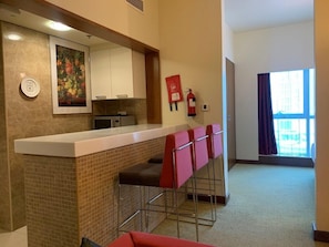 A modern kitchen with a white countertop, a microwave, and a fire extinguisher. There are four red bar stools and a painting on the wall. - Adagio Fujairah (Al-Fujairah)