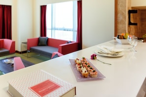 Habitación de hotel con una mesa de comedor lista para una comida, un sofá y una ventana con cortinas.