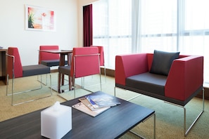 Quarto de hotel moderno com sofá, cadeiras, mesa e janela com cortinas.