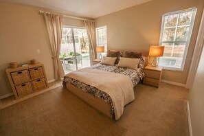 A bedroom with a large bed, a nightstand, a lamp, a window with blinds, and a sliding glass door leading to a balcony. - Southern Dunes Homes (Haines City)