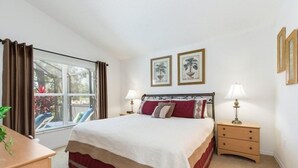 Un dormitorio con cama, mesitas de noche, una ventana con cortinas y dos cuadros en la pared.
