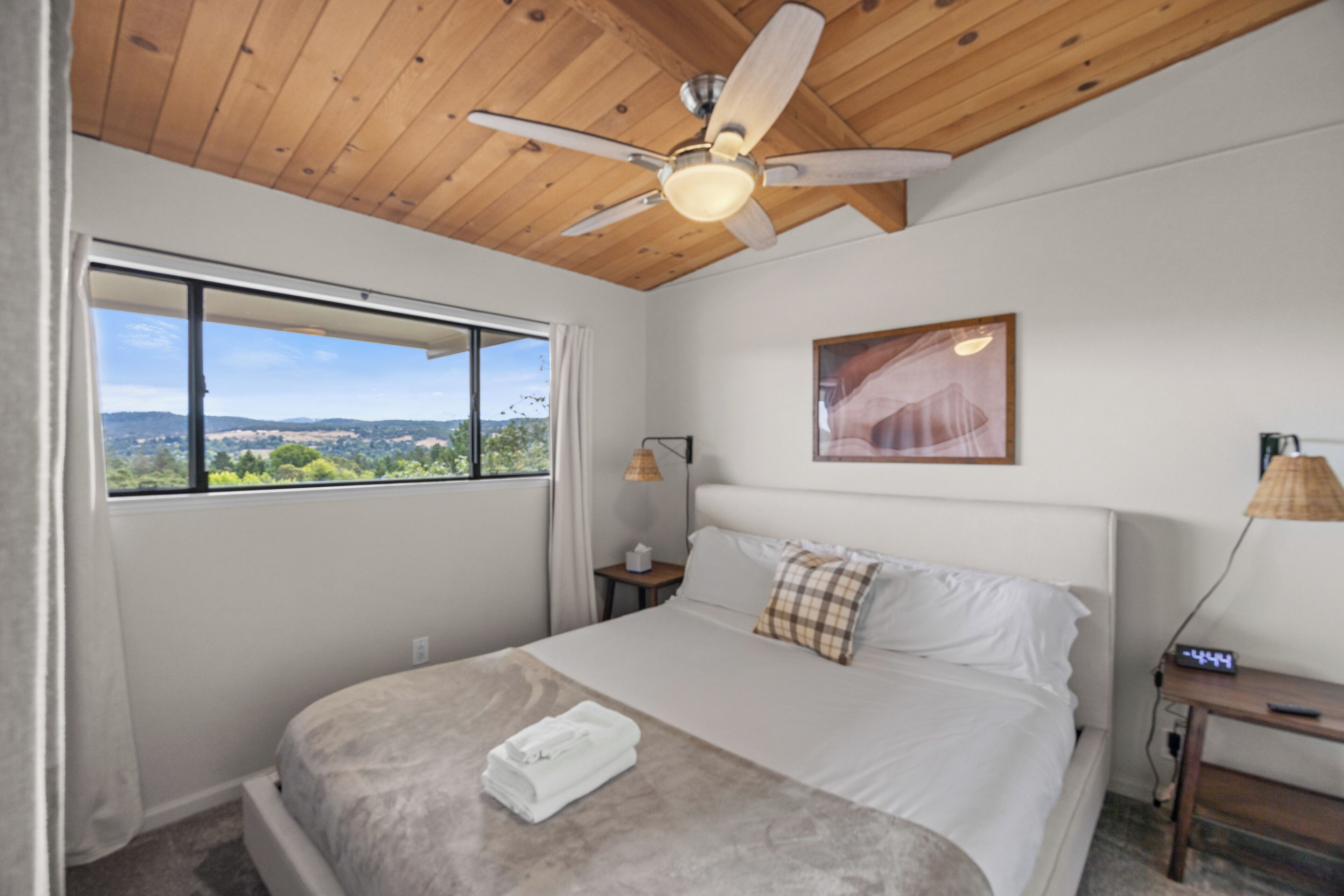 A bedroom with a bed, a ceiling fan, a window with a view, a bedside table, and a nightstand.