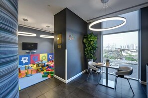 A modern office space with a children's play area, a large window with a city view, and a dining area with a plant. - Premier Inn Dubai Ibn Battuta Mall (Dubai)