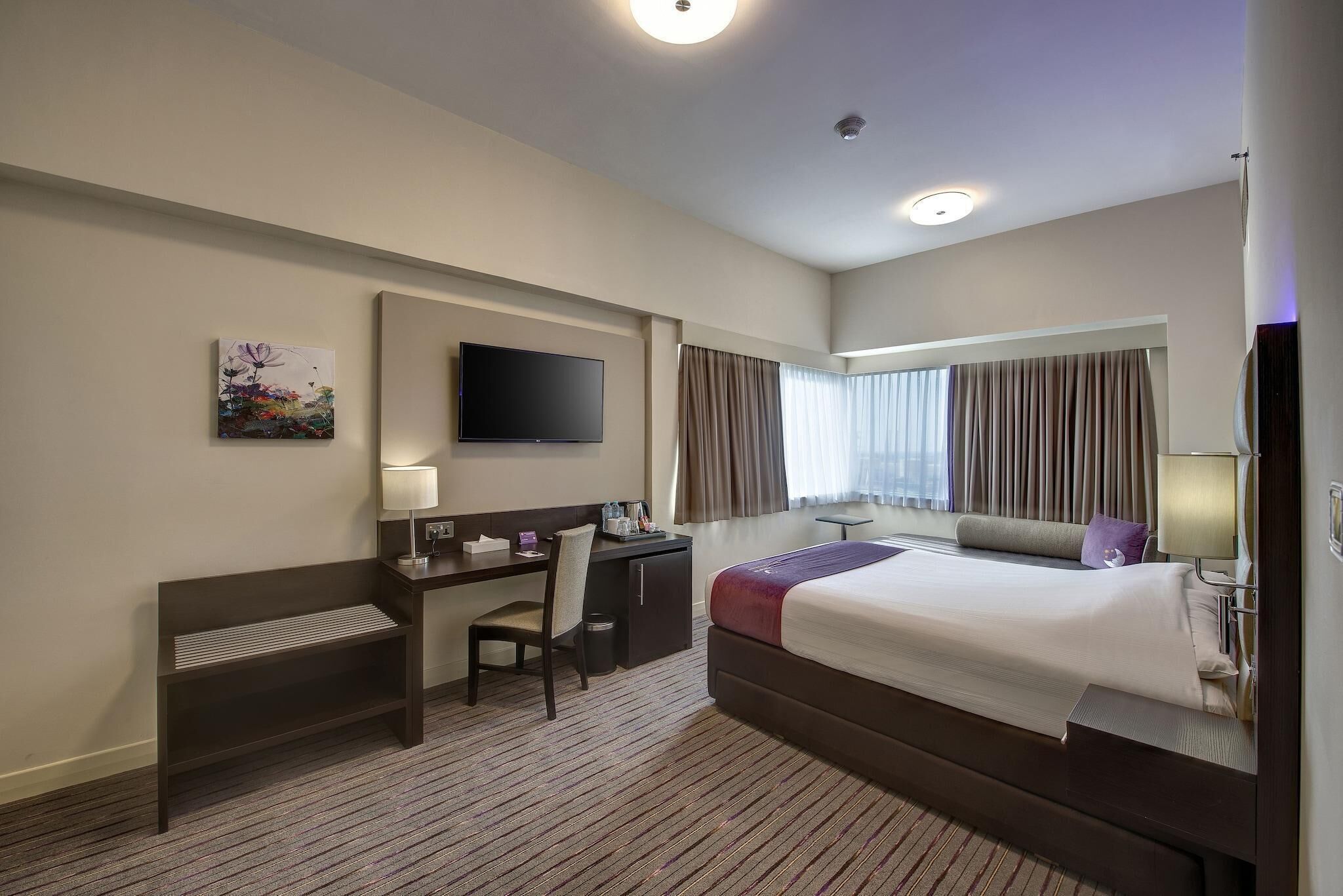 A hotel room with a bed, desk, chair, TV, and artwork on the wall.
