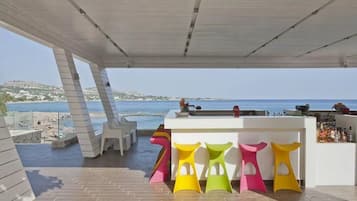 A modern outdoor seating area with a bar, colorful stools, and a view of the sea.