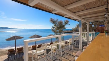 A beachside terrace with wooden flooring, white chairs, and umbrellas, overlooking the sea.