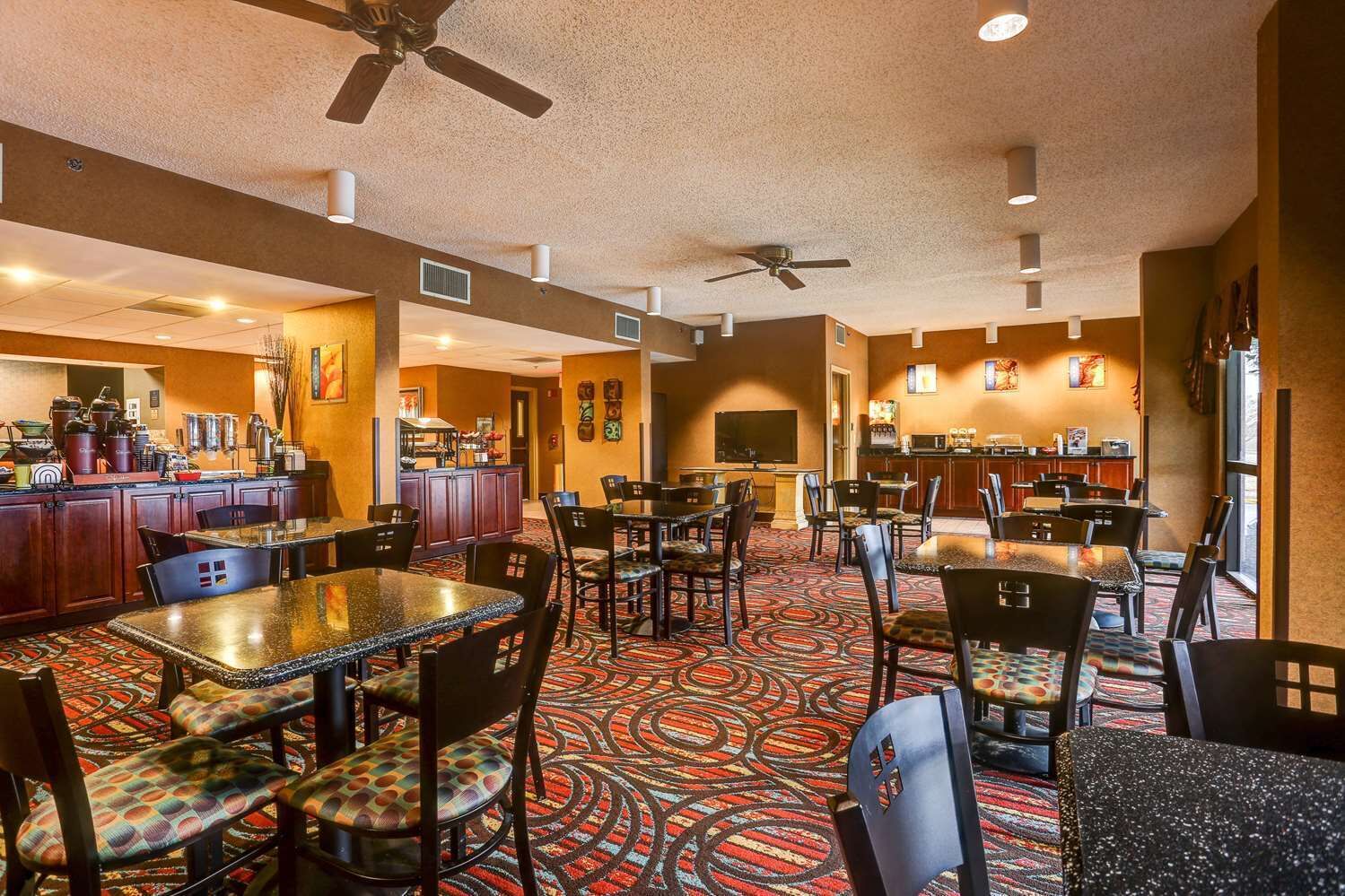 Zona pranzo dell'hotel con diversi tavoli e sedie, ventilatori a soffitto e un'area cucina sullo sfondo.