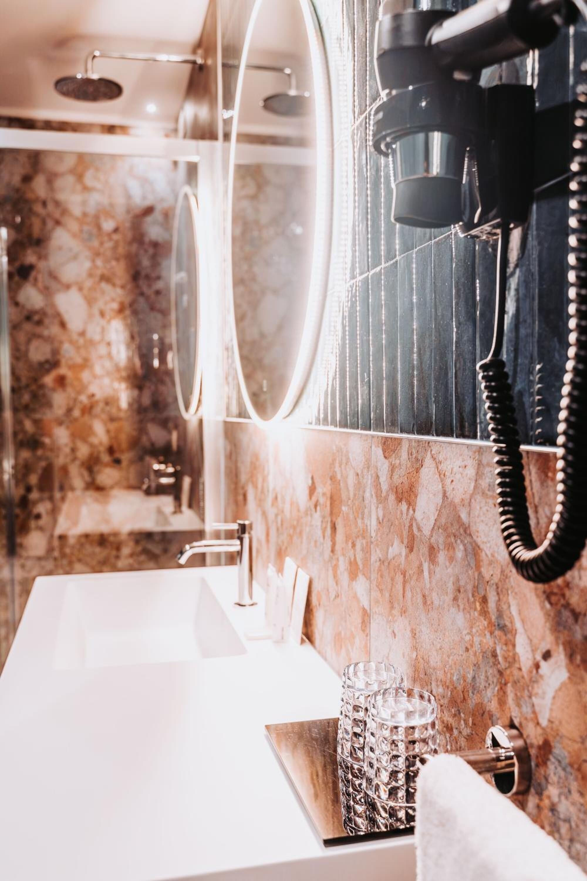 A bathroom with a marble wall, a white sink, a mirror with lights, and a hairdryer mounted on the wall.