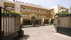 A hotel entrance with a sign reading 'AZALAI LE GRAND HOTEL,' a gated driveway, and a symmetrical facade with multiple windows.