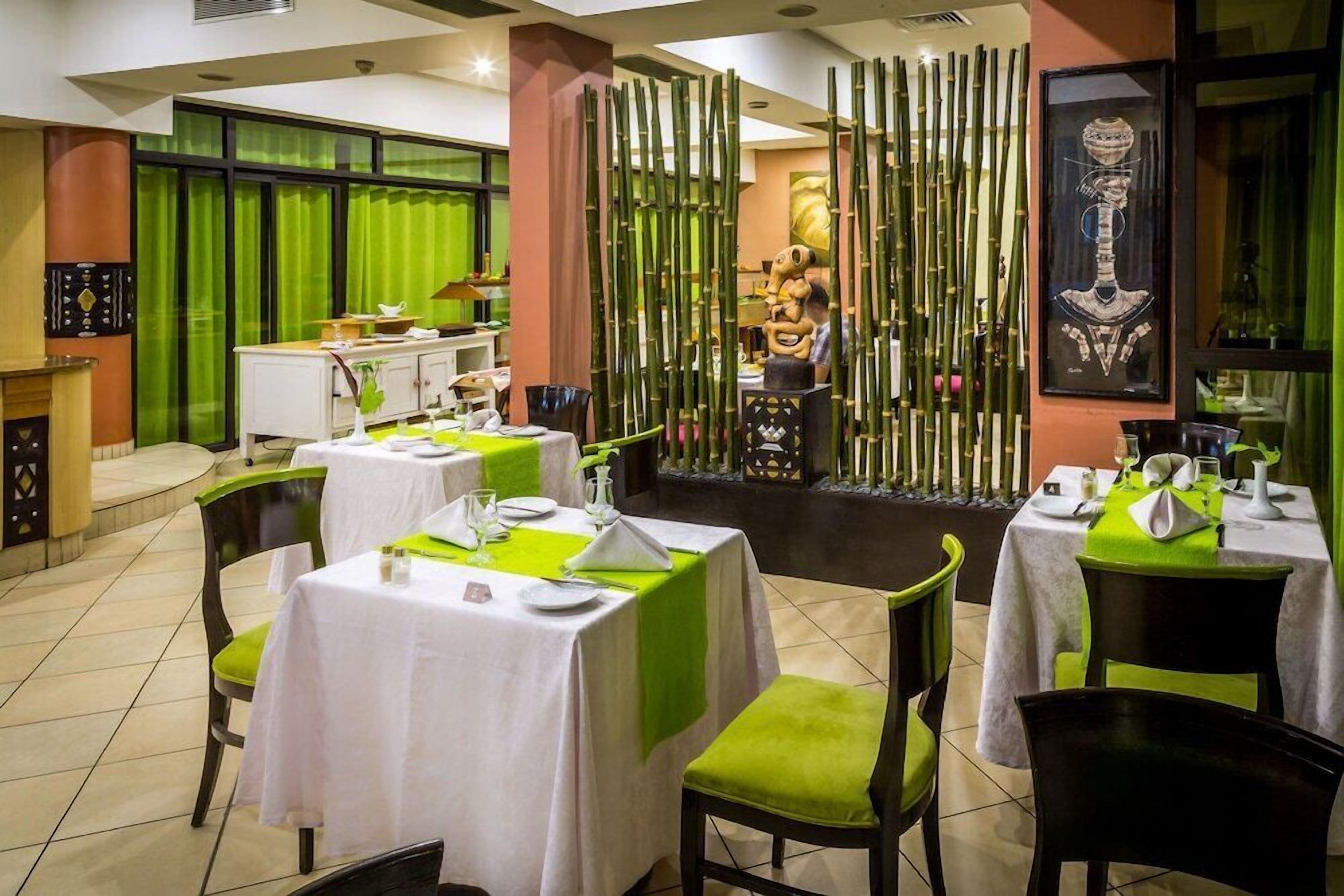 A restaurant with tables set for dining, a green curtain backdrop, and a bamboo screen in the background.