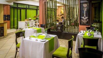 A restaurant with tables set for dining, a green curtain backdrop, and a bamboo screen in the background.