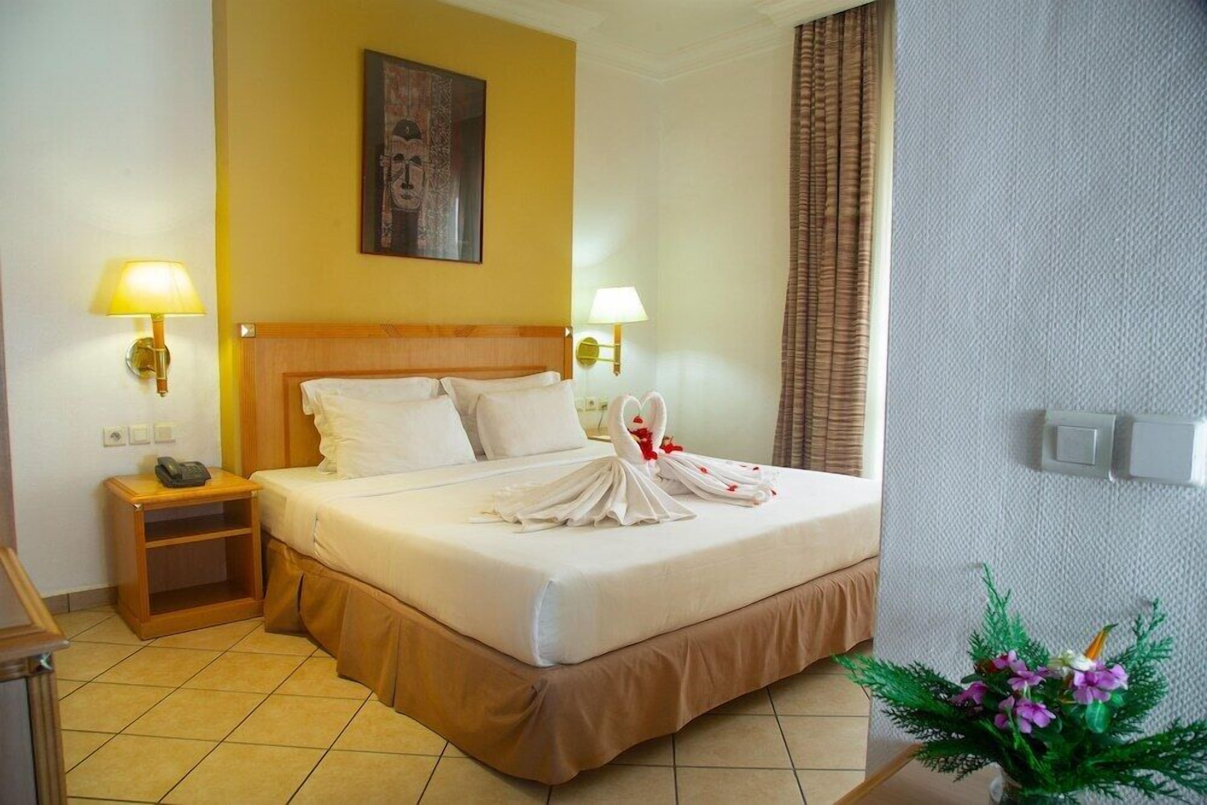 Une chambre d’hôtel avec un lit, deux lampes de chevet, une table de chevet, un téléphone, un tableau au mur et une plante en pot.
