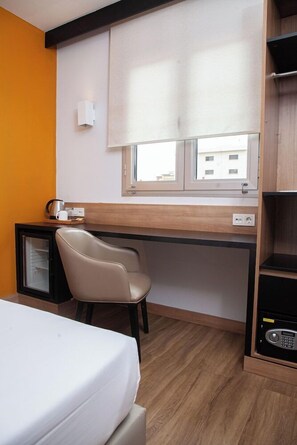 Une chambre d’hôtel comprenant un lit, un bureau avec une chaise, un petit réfrigérateur, une fenêtre avec des stores et une vue sur les bâtiments environnants.