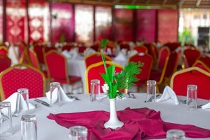 A banquet hall with tables set for dining, red chairs, and a white vase with greenery.