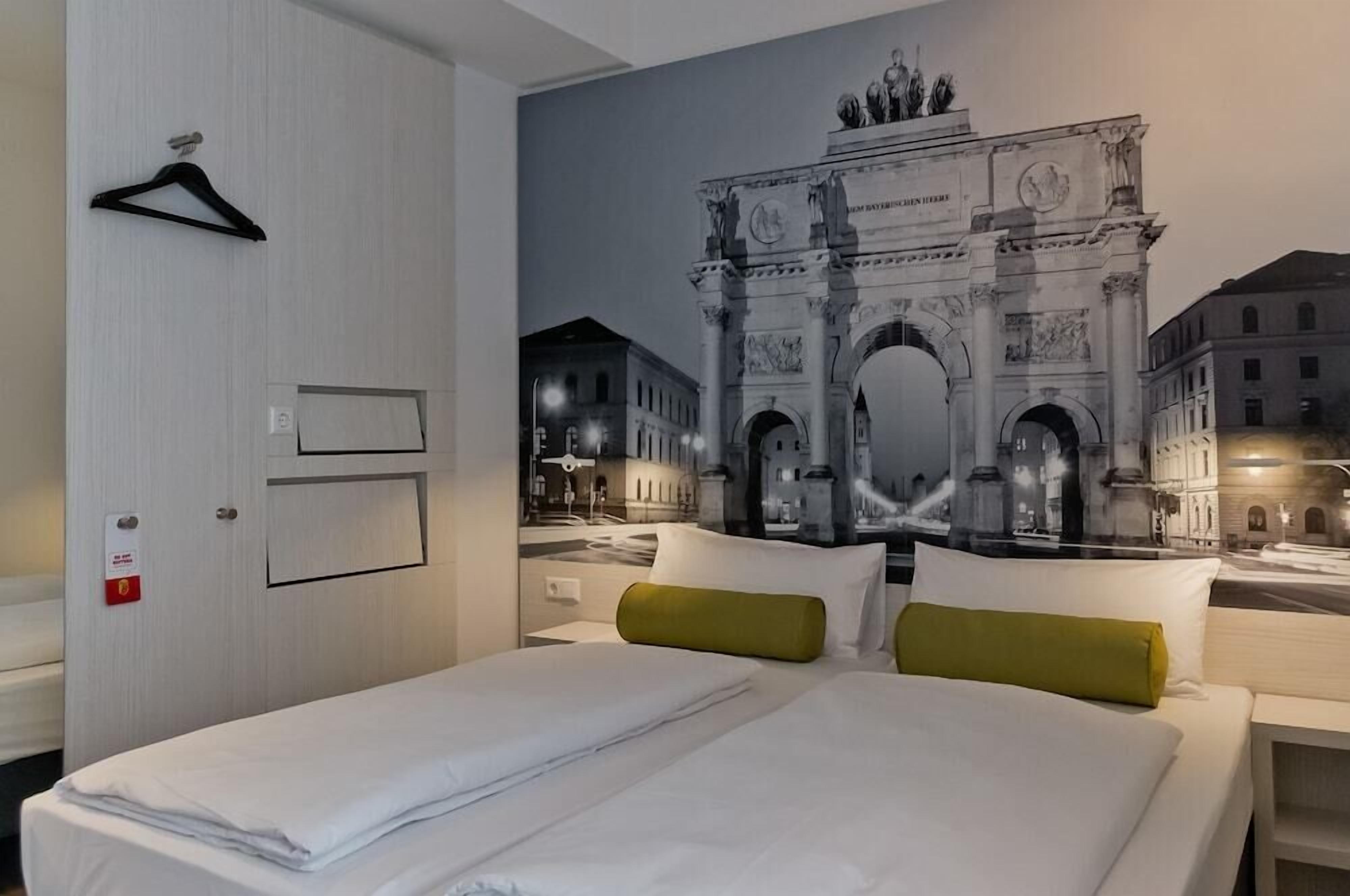 Ein Hotelzimmer mit Bett, Tür, Wandhänger und einem großformatigen Wandgemälde eines historischen Bogens.