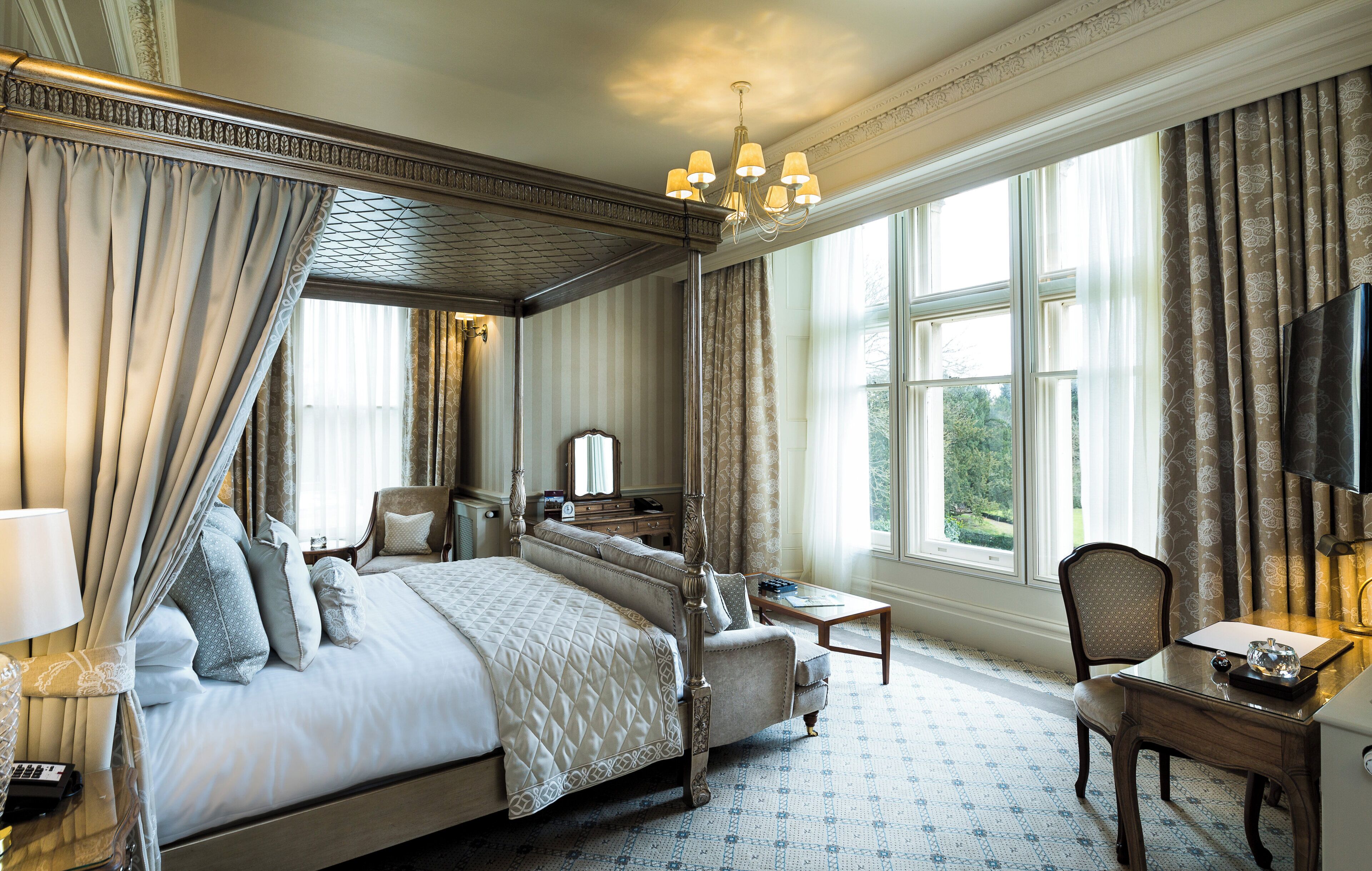 A four-poster bed with a canopy, a desk with a chair, a television, and a large window with curtains.