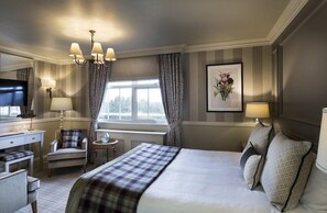 A hotel room with a bed, a desk, a chair, a television, a window with curtains, and a wall-mounted picture. - Down Hall Hotel Spa & Estate (Bishop's Stortford)