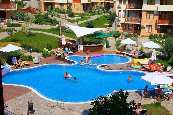 Una piscina con varias pistas, rodeada de sillas de descanso y sombrillas, ubicada en un patio con edificios al fondo.