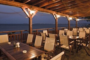 Un área de comedor al aire libre techada, con mesas y sillas de madera, con vista a un cuerpo de agua.