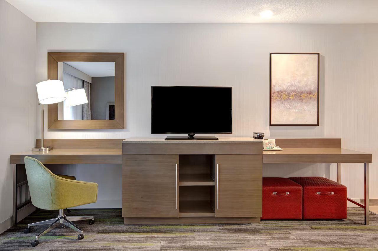 A hotel room with a desk, a television, a mirror, and a red suitcase.