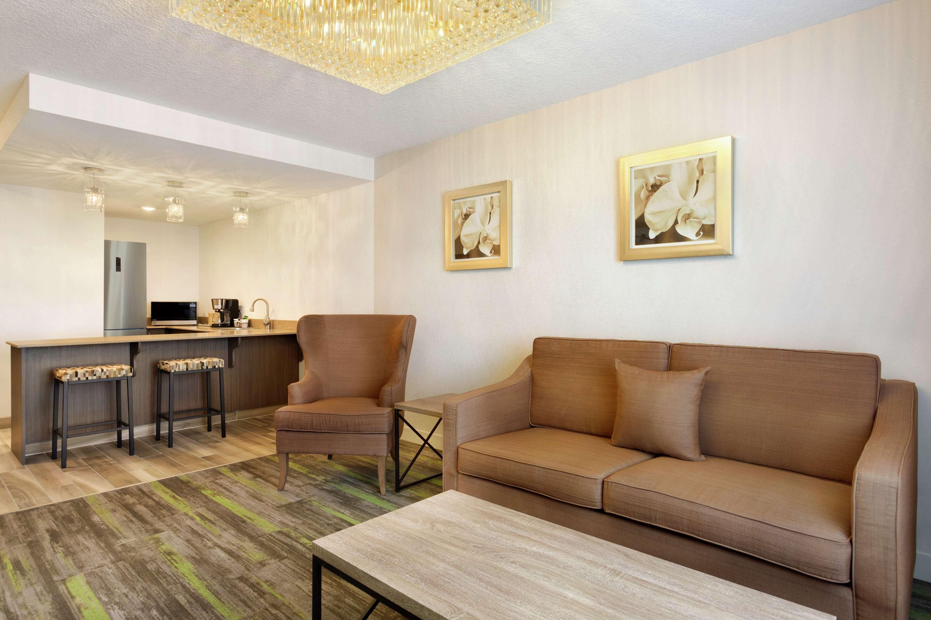 A modern living room with a sofa, armchair, and coffee table. There is a kitchen area with a refrigerator, microwave, and bar stools. Two framed pictures are on the wall.