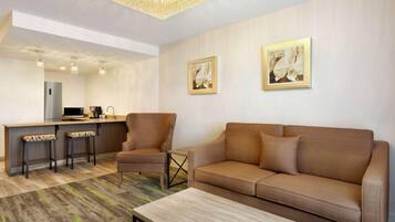 A modern living room with a sofa, armchair, and coffee table. There is a kitchen area with a refrigerator, microwave, and bar stools. Two framed pictures are on the wall.