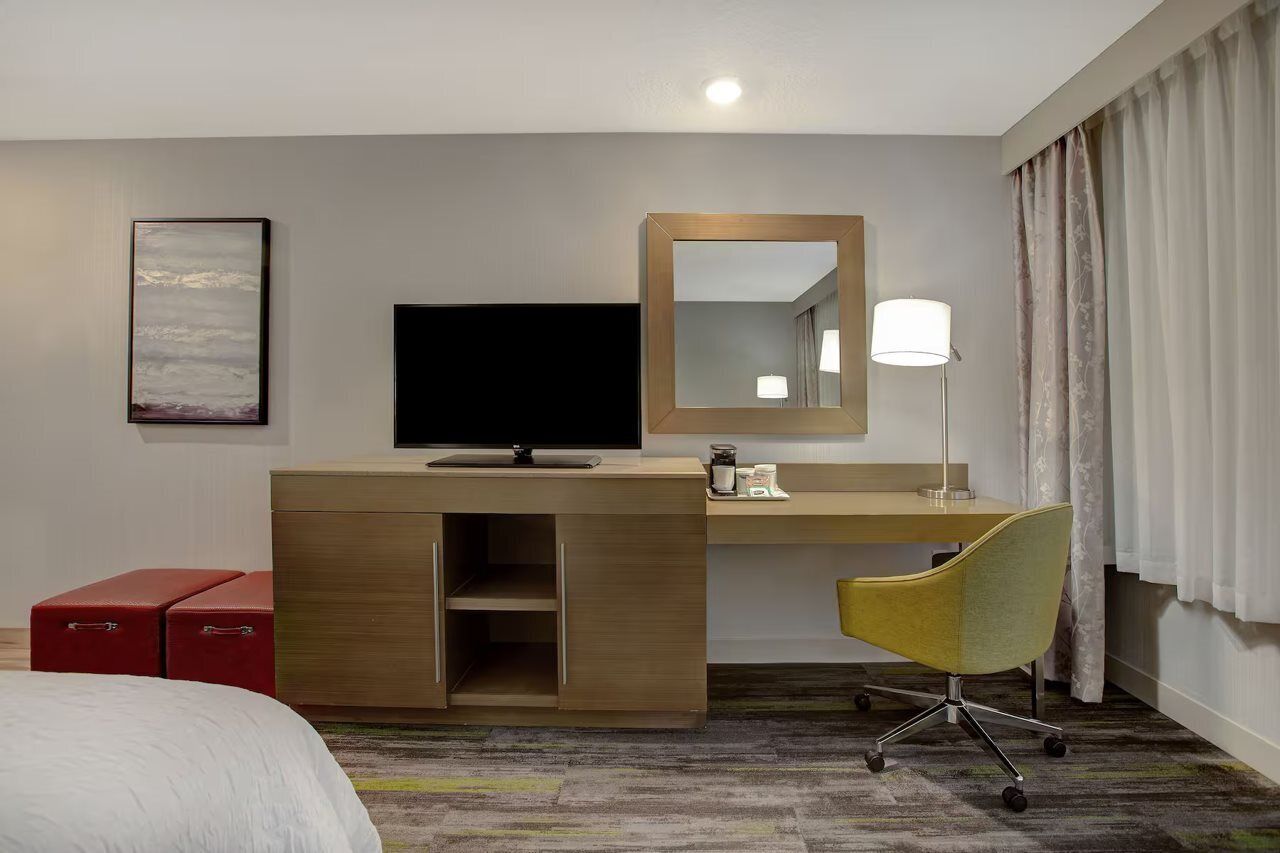 A hotel room with a bed, a desk with a flat-screen TV, a mirror, a lamp, and a red storage unit.