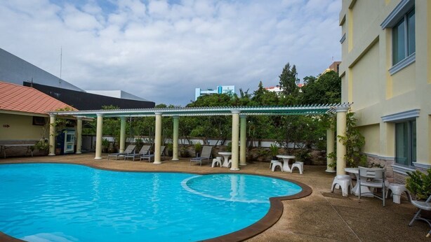 A swimming pool with a curved edge, surrounded by lounge chairs and small tables, with a covered seating area and a building in the background. - Eco Hostel (Hua Hin)