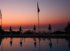 Blick auf ein Schwimmbecken mit Liegestühlen und Sonnenschirmen, eine Fahnenmasten und ein Sonnenuntergang über dem Wasser.