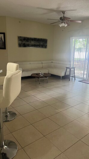 Una sala de estar con sofá blanco, una mesa de centro, ventilador de techo y un ventanal con persianas.