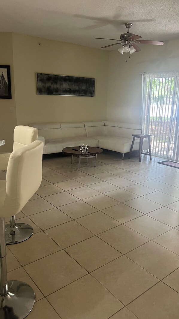 A living room with a white sofa, a coffee table, a ceiling fan, and a large window with blinds.