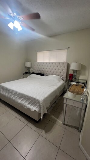 A bedroom with a bed, a nightstand, a bedside table, a ceiling fan, and a window with blinds.