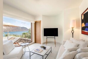 A modern living room with a large window, a flat-screen TV, a sofa, a coffee table, and a lamp. - Phea Blue Palace (Agios Nikolaos)