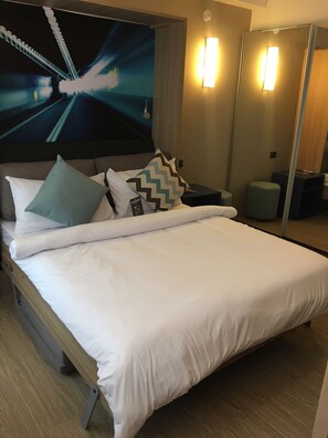 A hotel room with a large bed, a headboard with a cityscape mural, bedside lamps, and a small seating area.